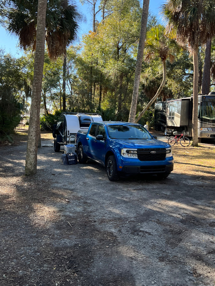 Blue truck with rv rental camper at a sunny campsite. Larger motorhomes and palm trees in this serene camping setting.