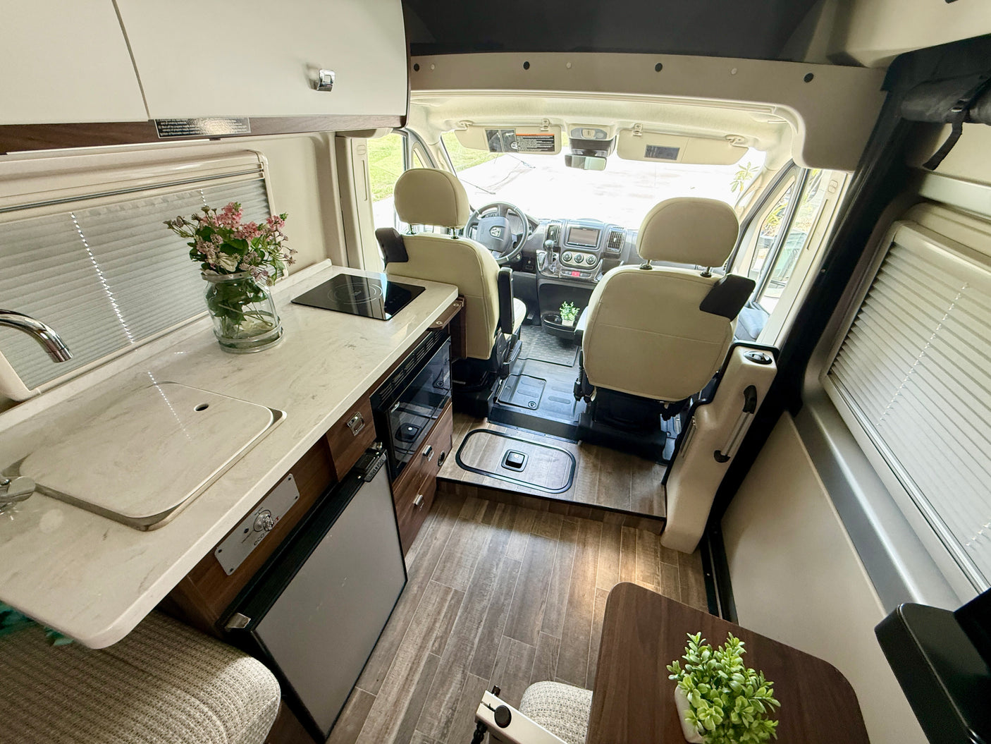 Interior of a modern rv rental: shows kitchen counter with sink & stove, fridge, wood flooring, and driver's cabin.