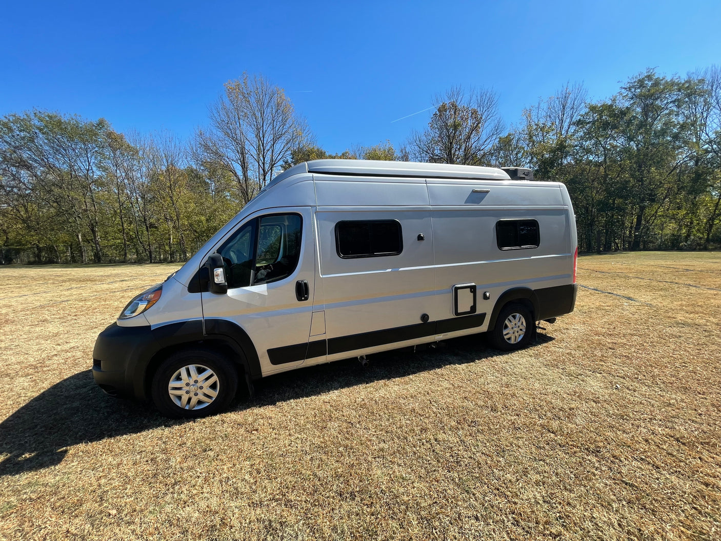 This silver camper van, a perfect RV rental, is parked on a sunny dry field with trees & a clear blue sky. Explore the outdoors!