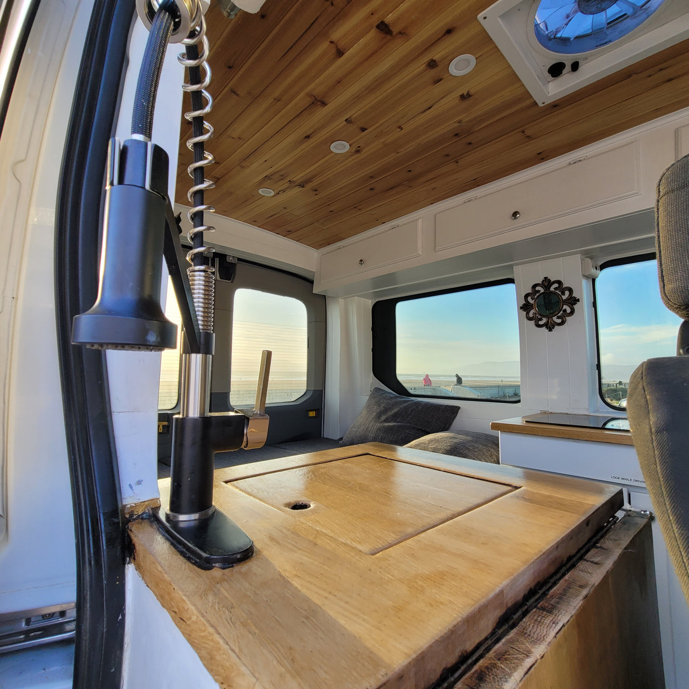 This RV rental interior shows a cozy converted van with a kitchen area, wooden ceiling, and a stunning beach view from the windows.