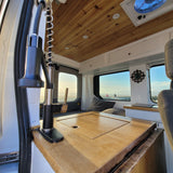 This RV rental interior shows a cozy converted van with a kitchen area, wooden ceiling, and a stunning beach view from the windows.