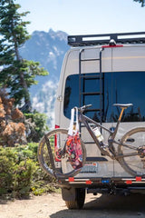 RV rental van, ready for adventure with a mountain bike on its rear rack, ladder to roof rack, and scenic mountains.