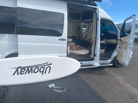 Experience van life! This white camper van, an ideal RV rental, shows a sleeping dog inside & a paddleboard outside.