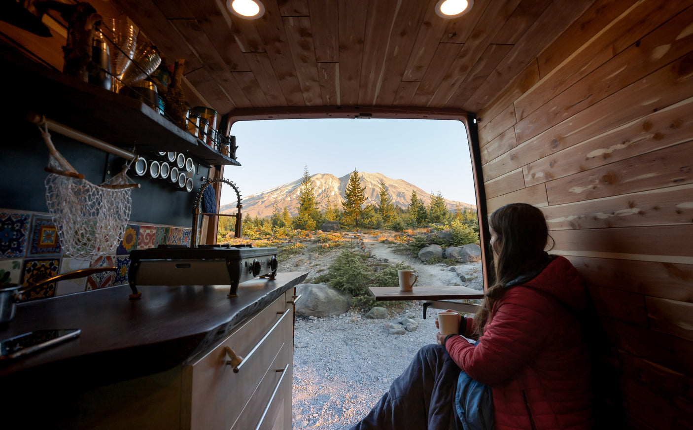 Cozy RV rental: Woman sips coffee from wood-paneled van, enjoying mountain & forest view from open rear.