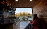 Cozy RV rental: Woman sips coffee from wood-paneled van, enjoying mountain & forest view from open rear.