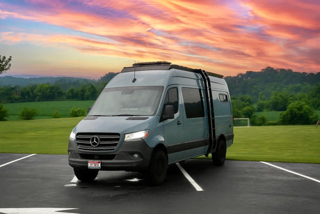 Grey Mercedes Sprinter RV rental with roof rack & ladder, parked in a scenic landscape at sunset.