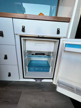 RV rental's compact refrigerator in a camper van, featuring an open door revealing a freezer, wire shelf, and blue drawers.