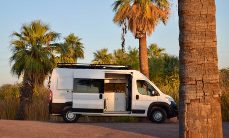 White camper van RV rental parked on a dirt path with its side door open, revealing a modern kitchen. Palm trees surround it.