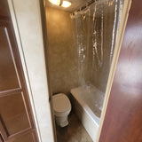 RV rental bathroom with a toilet on the left and a bathtub/shower with a clear curtain on the right, lit by an overhead fixture.