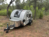 Compact teardrop RV rental, grey with black trim, parked in a dirt clearing among green forest trees for camping.