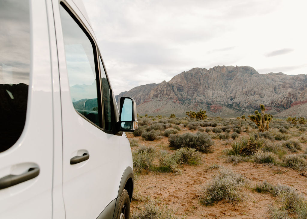 Scenic rv rental adventure. White van parked in a desert with rugged mountains, shrubs, and Joshua trees under cloudy skies.