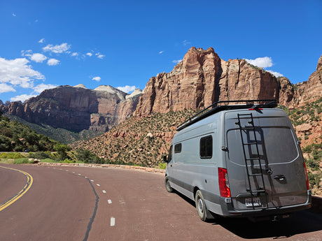 An RV rental camper van parked on a scenic road amidst Zion National Park's towering red rock mountains under a blue sky.
