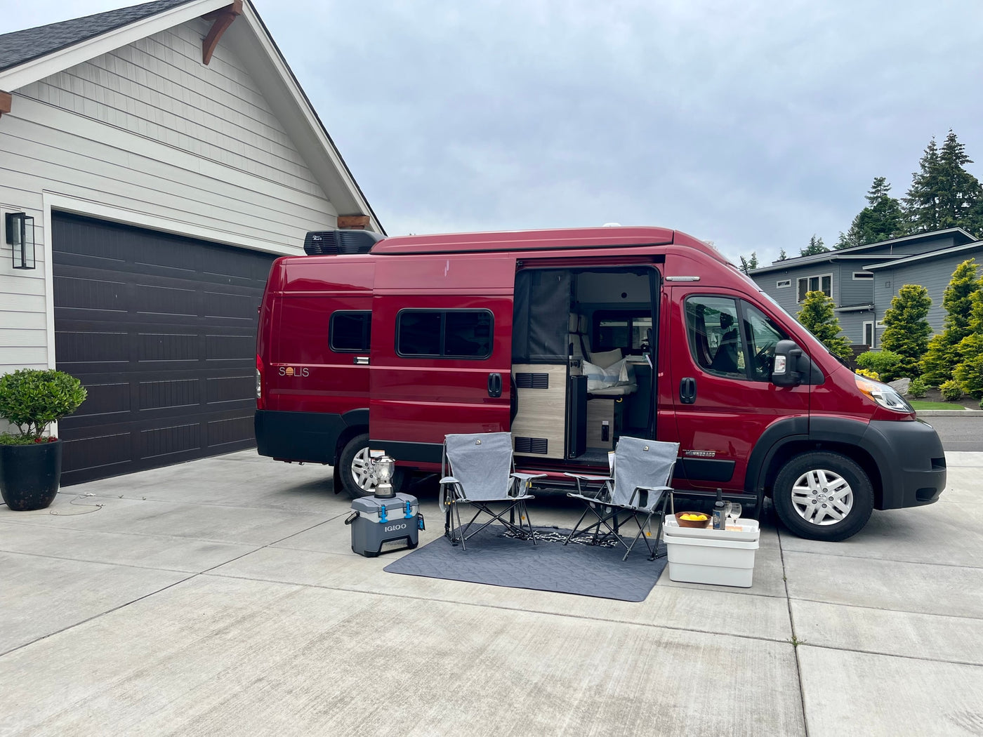 Relaxing by a red Winnebago Solis rv rental: a cozy outdoor setup with chairs, cooler, and open camper van door in a driveway. — photo 11