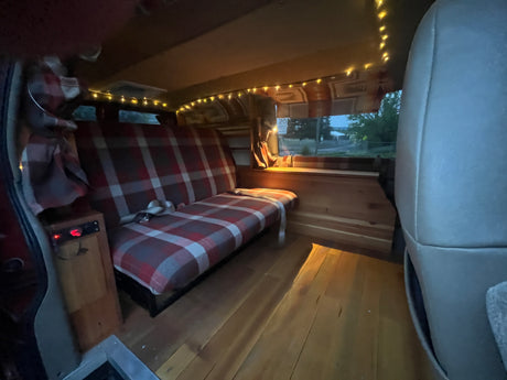 Step inside this cozy RV rental. Plaid bench seat, wooden interior, & ambient string lights create a perfect camper van setup.