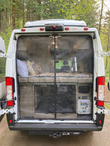 Open back of an rv rental camper van, revealing a bed, storage, and utility panel behind a protective bug screen. Ideal for camping.