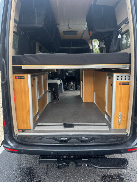 RV rental camper van interior: rear view showcasing a raised bed, modular Trail Kitchens cabinets, and overhead storage.