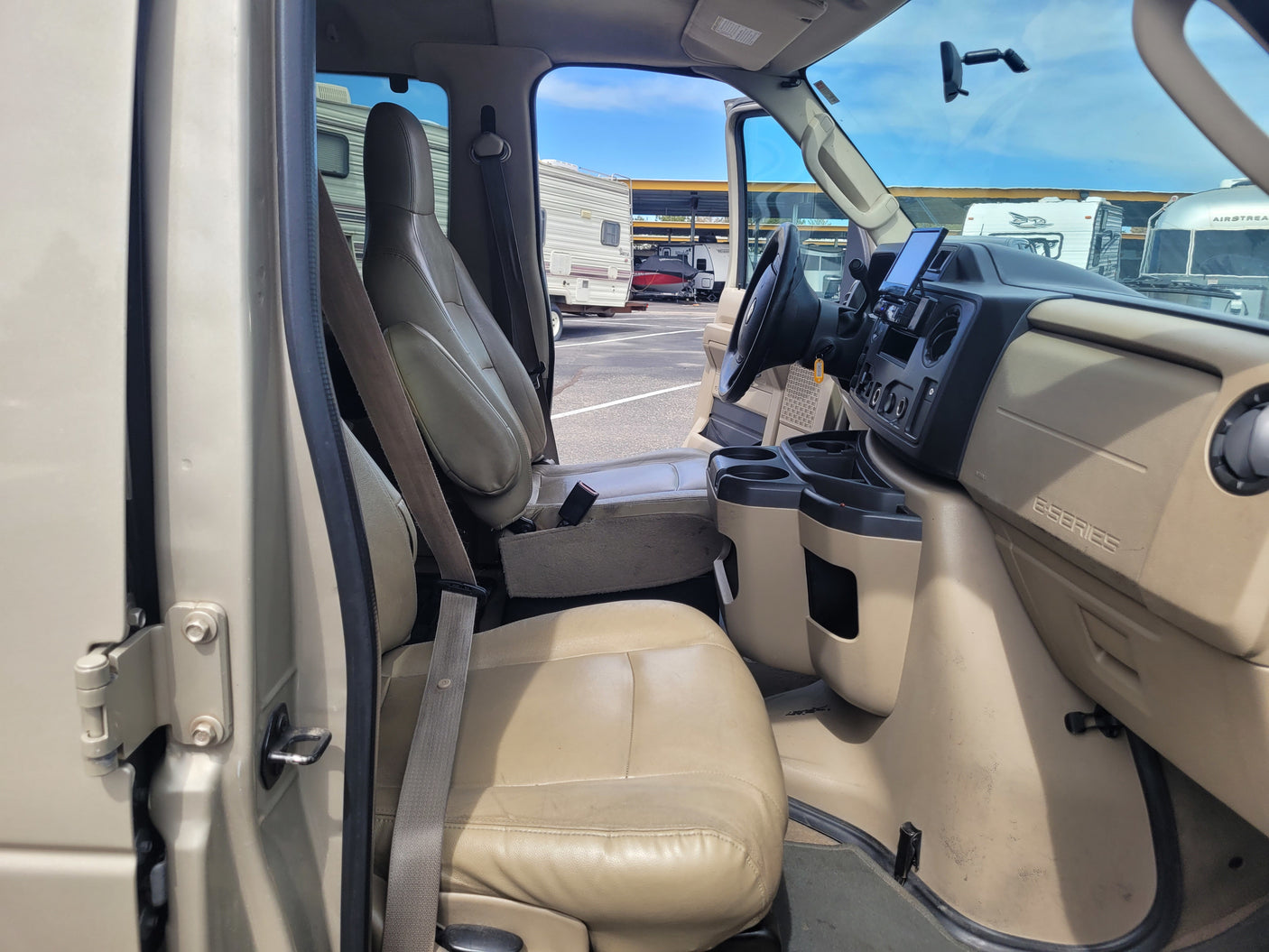 Inside a Ford E-Series RV rental. Driver's cockpit with steering wheel, dashboard screen, and passenger seat visible. RVs are outside.
