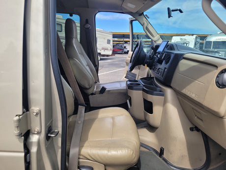 Inside a Ford E-Series RV rental. Driver's cockpit with steering wheel, dashboard screen, and passenger seat visible. RVs are outside.