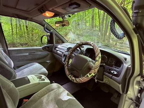 Inside an RV rental: driver's area with custom steering wheel, dashboard, and lush forest view. Perfect for nature adventures.