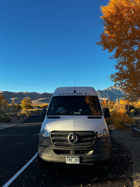 A white Mercedes Sprinter rv rental van among vibrant fall leaves, with distant sand dunes and mountains under a clear blue sky.