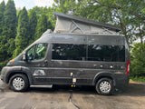 Dark grey pop-top camper van (RV rental) with adventure decals, parked by lush trees on asphalt.