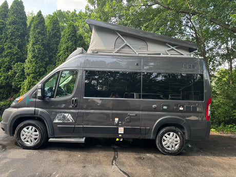 Dark grey pop-top camper van (RV rental) with adventure decals, parked by lush trees on asphalt.