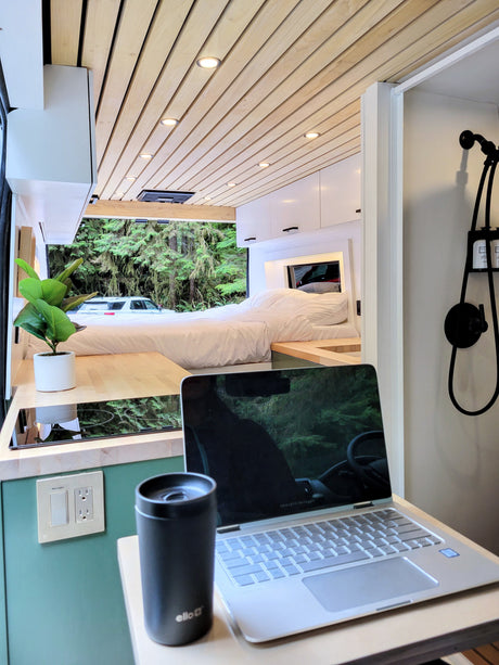 Bright RV rental interior: cozy bed, modern kitchen, and a laptop work setup with a scenic forest view outside.