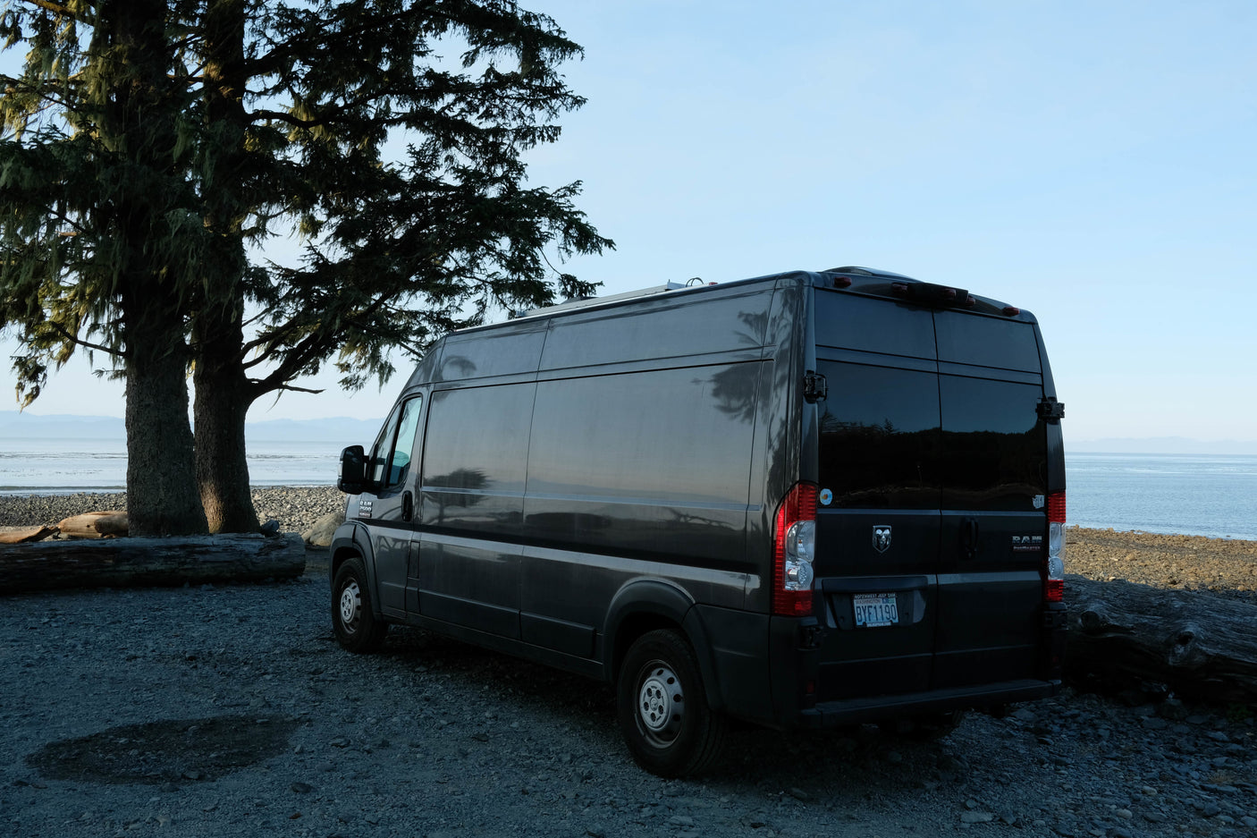 Dark grey Ram ProMaster camper van, possibly an RV rental, parked on a rocky beach by the ocean with large trees.