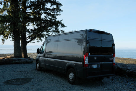 Dark grey Ram ProMaster camper van, possibly an RV rental, parked on a rocky beach by the ocean with large trees.