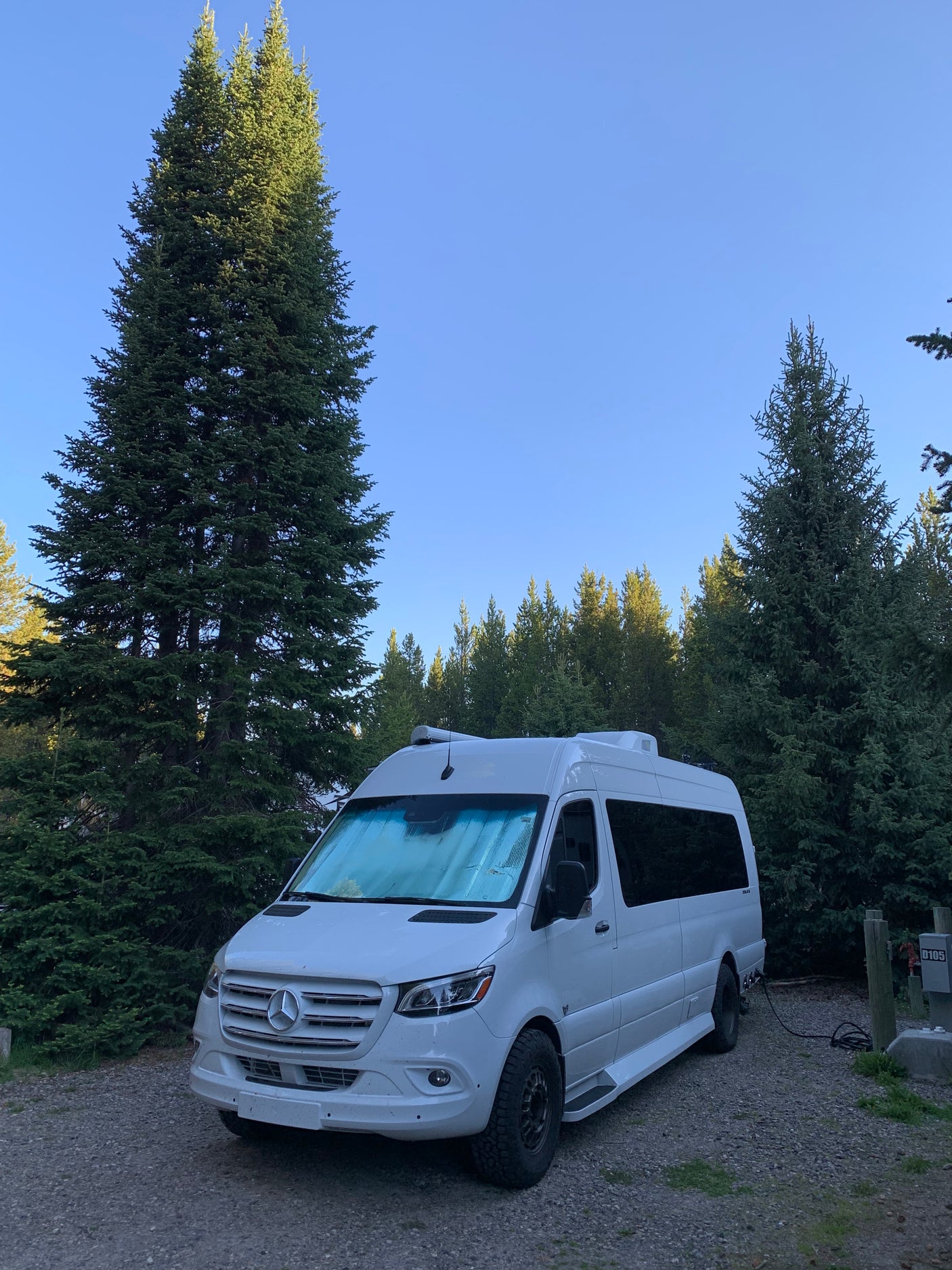 This white Mercedes Sprinter RV rental is parked at a forested campsite, hooked to power for a cozy stay.