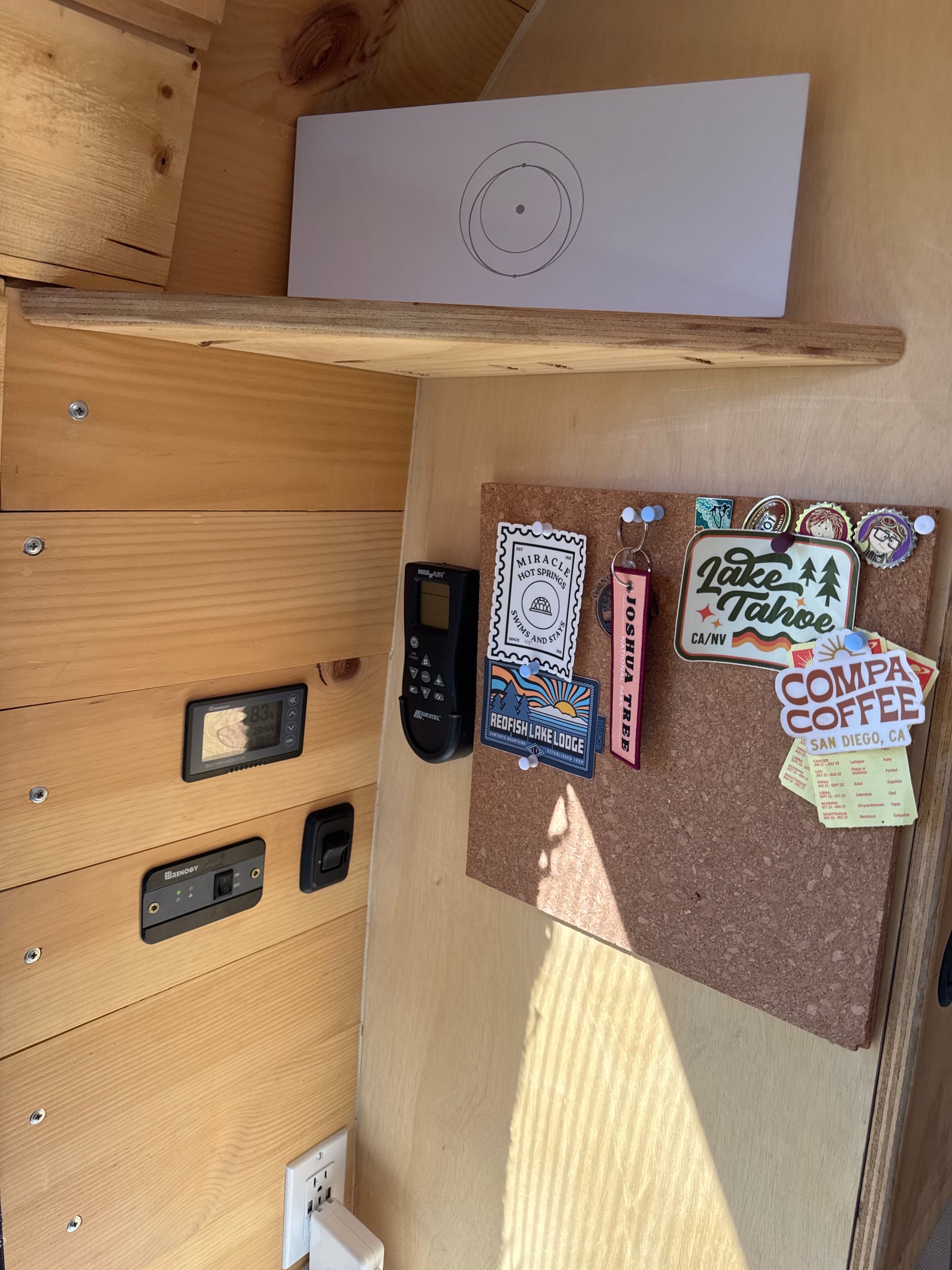 Cozy RV rental interior with wood paneling, travel stickers on a corkboard, battery monitor, and fan remote.