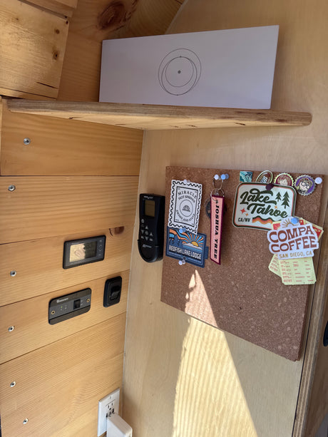 Cozy RV rental interior with wood paneling, travel stickers on a corkboard, battery monitor, and fan remote.