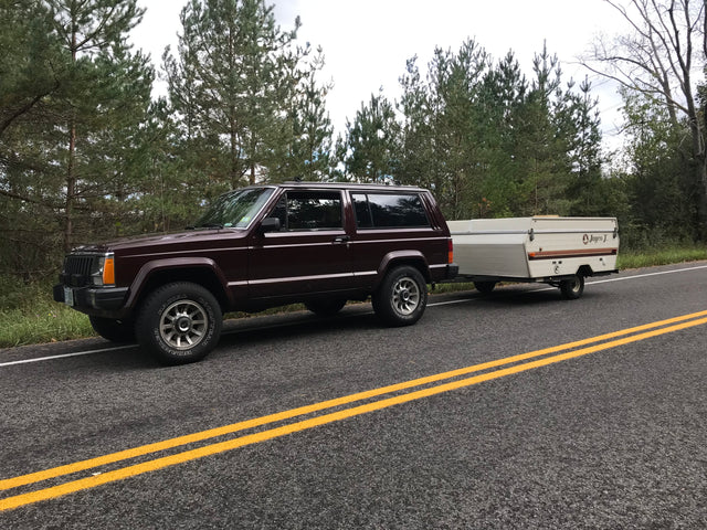 Dark red Jeep Cherokee towing a white Jayco J pop-up camper on a roadside. Great for RV rental camping!