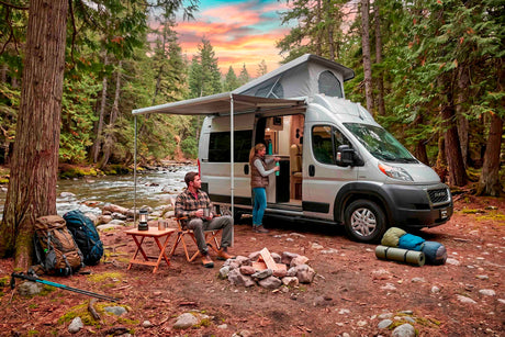 Enjoy your RV rental adventure! A couple relaxes by a river with their camper van, campfire, and hiking gear at sunset.