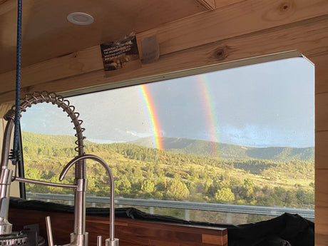 A stunning double rainbow over lush mountains, viewed from an rv rental window showing a modern kitchen faucet & cozy wooden interior.
