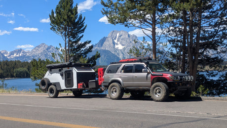 Modified SUV towing an XTR off-road camper by a lake & snow-capped mountains. Consider this RV rental for adventure!