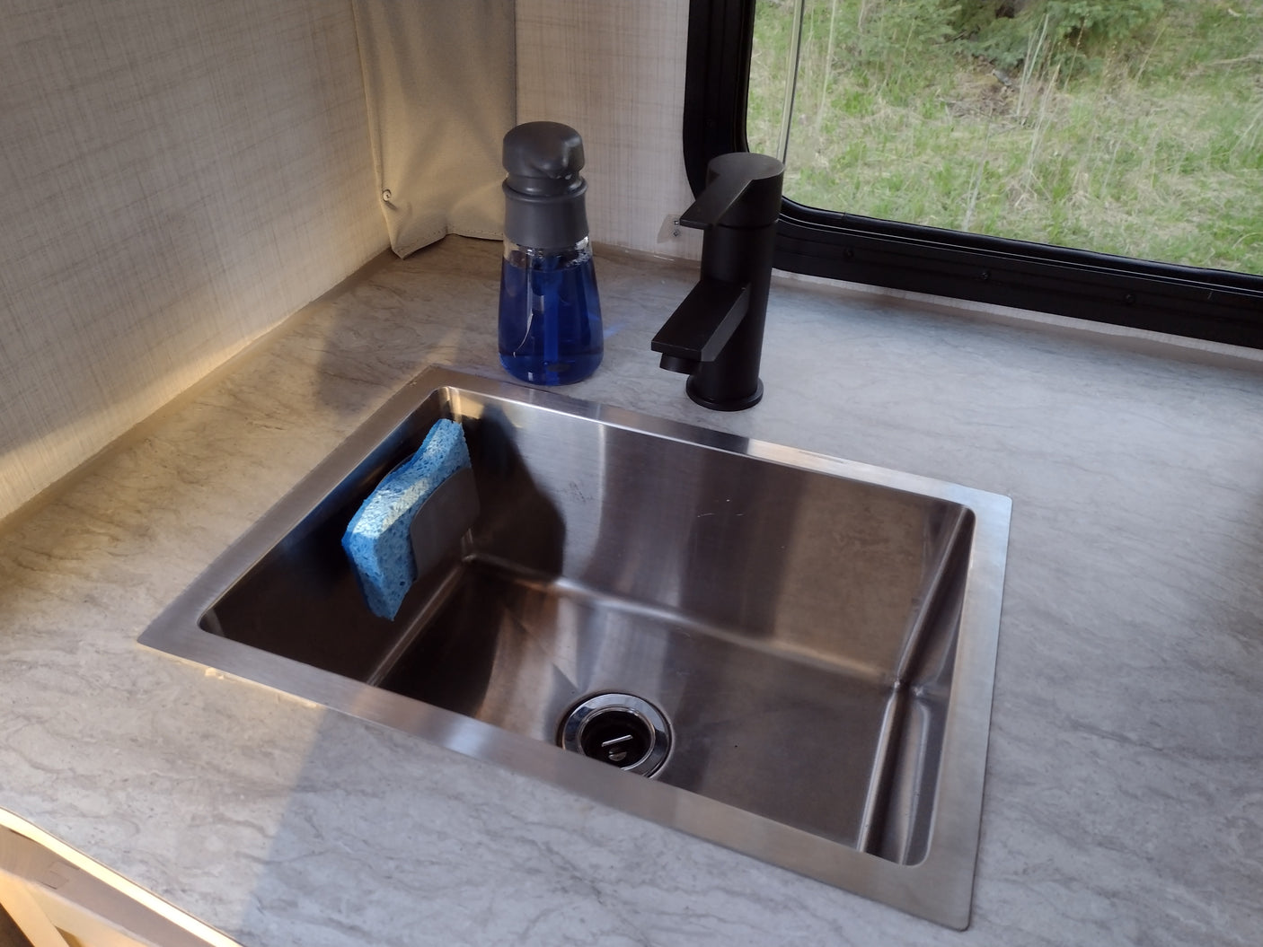 RV rental kitchen counter with stainless steel sink, blue sponge, black faucet, blue soap bottle, and a window.