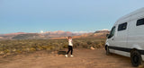 Explore the desert! A woman smiles by her RV rental with stunning mountains and a bright moon in the background.