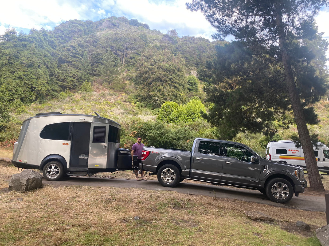 A man stands with his truck and a unique RV rental travel trailer at a beautiful forested campsite, near another camper van. — photo 9