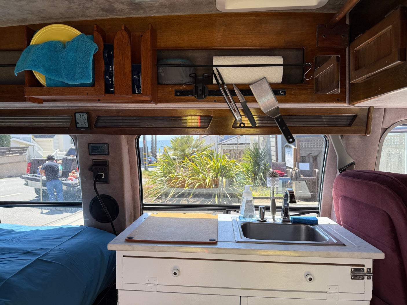 Interior of an rv rental camper van, showing a compact kitchen with sink, utensils, and bed, plus digital displays and an outdoor view.