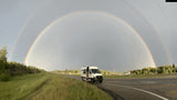 Stunning double rainbow arcs over a white RV rental van parked on a rural highway after a rain shower.
