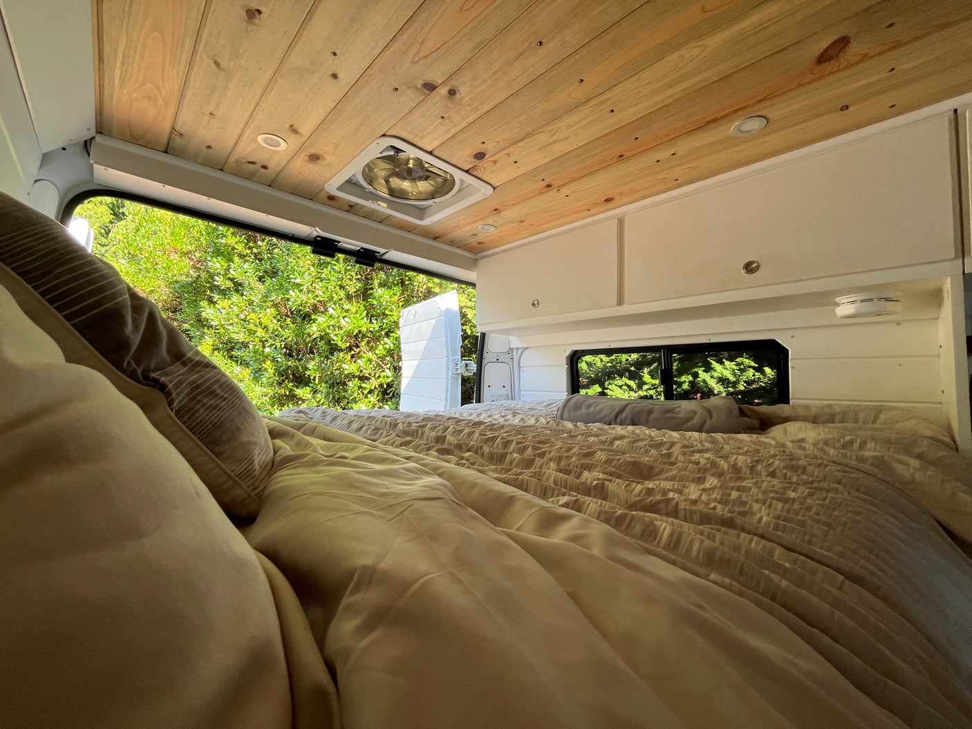 Cozy RV rental camper van interior. Features a comfy bed, wood ceiling, and serene green nature view.
