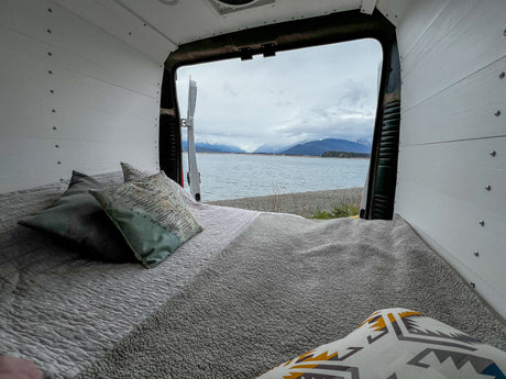 Experience nature from your cozy RV rental bed! Open van doors reveal a stunning Alaskan landscape: water, mountains, and sky.