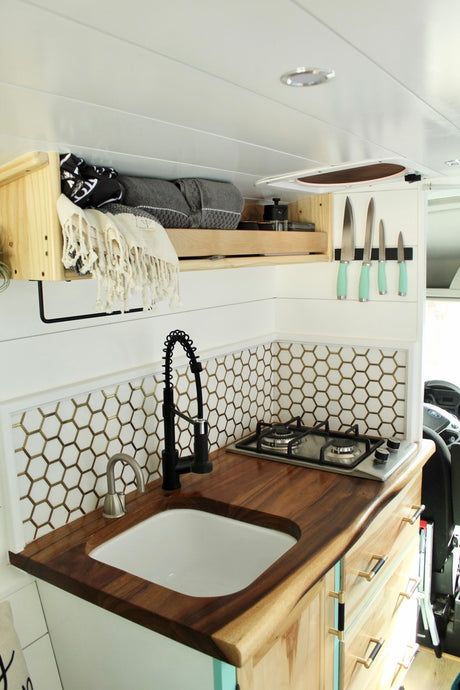 This compact RV rental kitchen features wood counters, a sink, stove, hex tile backsplash, magnetic knife strip, and overhead storage.