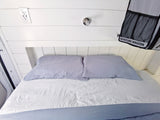 Comfortable RV rental bed setup with white shiplap, power outlet, and "ADVENTURE WAGON" storage bag.