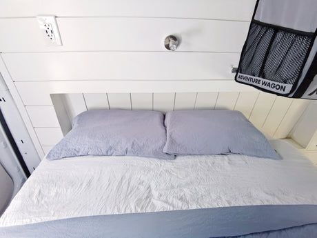 Comfortable RV rental bed setup with white shiplap, power outlet, and "ADVENTURE WAGON" storage bag.