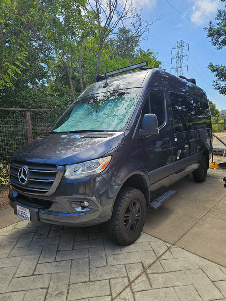 Dark grey Mercedes Sprinter van, an RV rental, with large tires & roof light bar, parked on pavers.