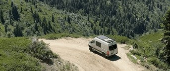 An RV rental camper van drives a winding dirt road through a densely forested mountain landscape.