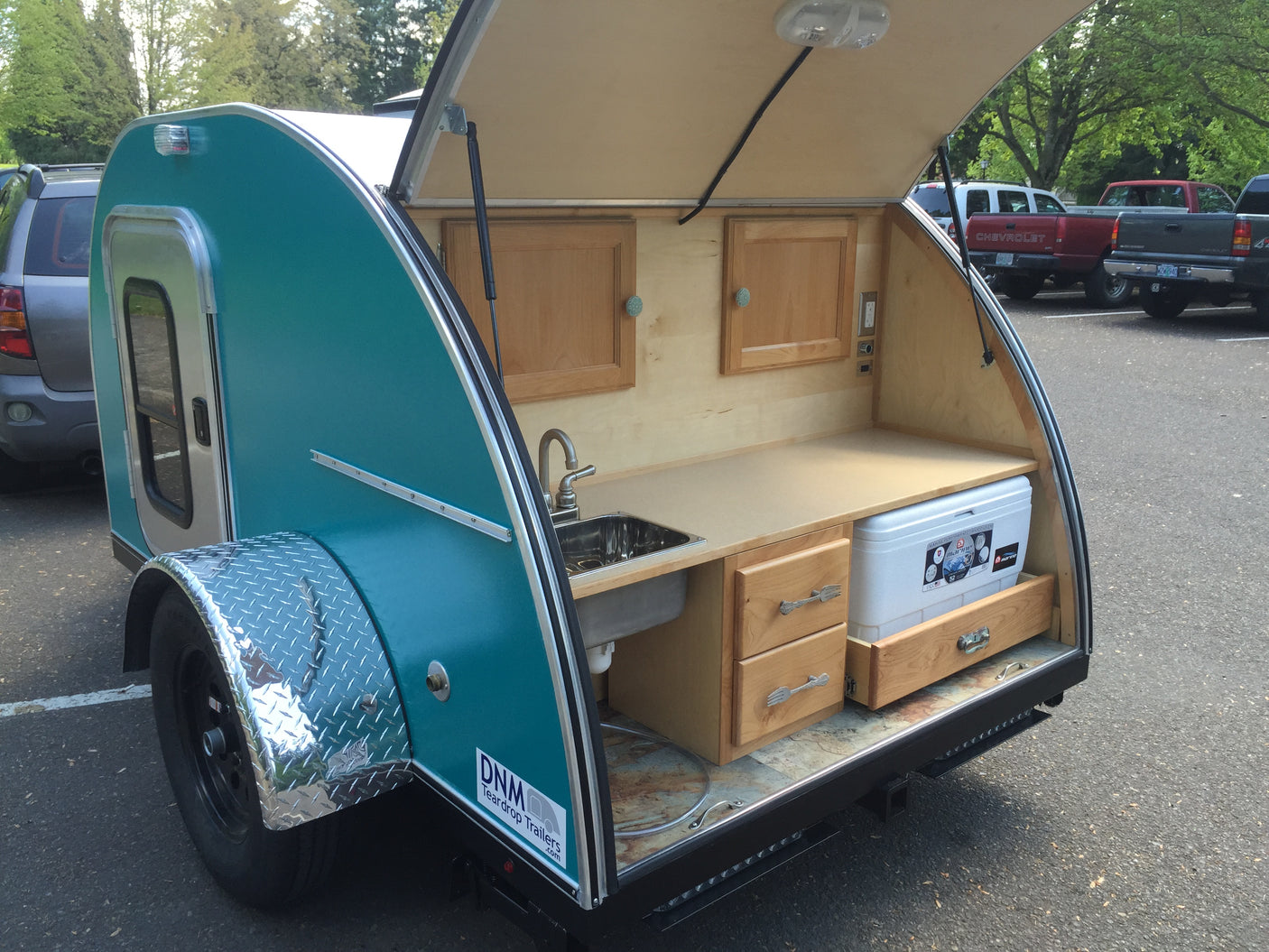 This teal teardrop RV rental showcases its outdoor kitchen galley with a sink, countertop, wooden cabinets, and cooler.