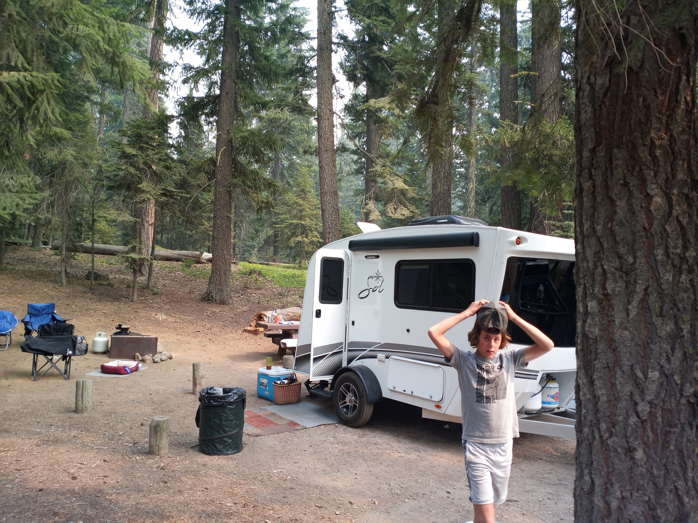 A boy at a forest campsite with a small RV rental, chairs, and fire pit under tall trees.